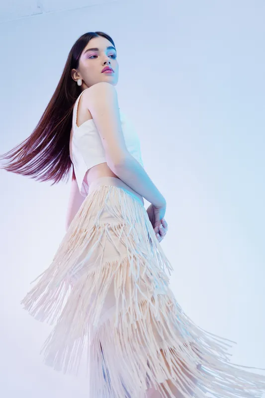 Young woman in stylish white top and beige fringed skirt poses confidently against a light background. Concept of woman beauty fashion, elegance, and modern style.