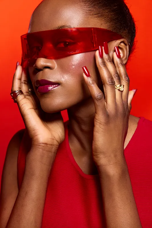Stylish dark skinned African woman with glossy makeup and red nails poses wearing red transparent glasses and matching outfit on vibrant red background. Concept of fashion and beauty.