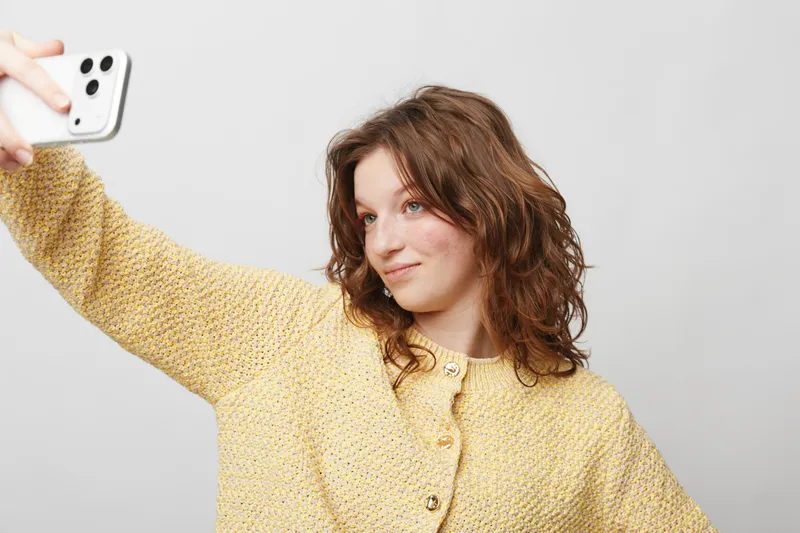 Young woman with curly hair wearing yellow sweater takes selfie with smartphone on white background. Concept of social media, self-portrait, casual style, and modern technology usage.