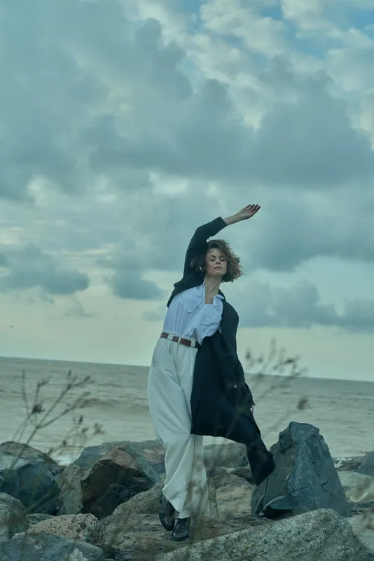 A stylish woman strikes a dynamic pose on a rocky shoreline under an overcast sky, wearing white pants, a belt, blazer, and blouse to convey movement, fashion, and a coastal mood.