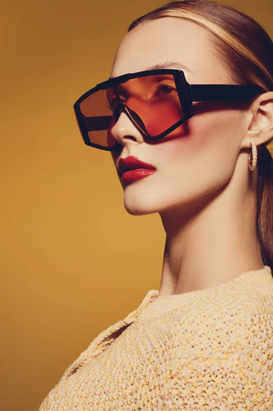 Blonde woman wearing large geometric sunglasses and red lipstick poses against yellow background. Fashion portrait with stylish makeup and trendy accessories in warm studio lighting.