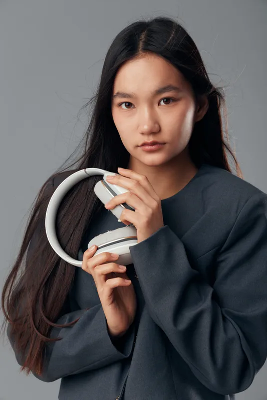 Young woman with long hair holding headphones close to her chest wearing dark gray jacket on gray background in studio. Casual style and thoughtful facial expression closeup.