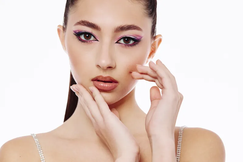 Young woman with creative fashion makeup poses with hands near face on a white background emphasizing glamour style and beauty trends in studio light.