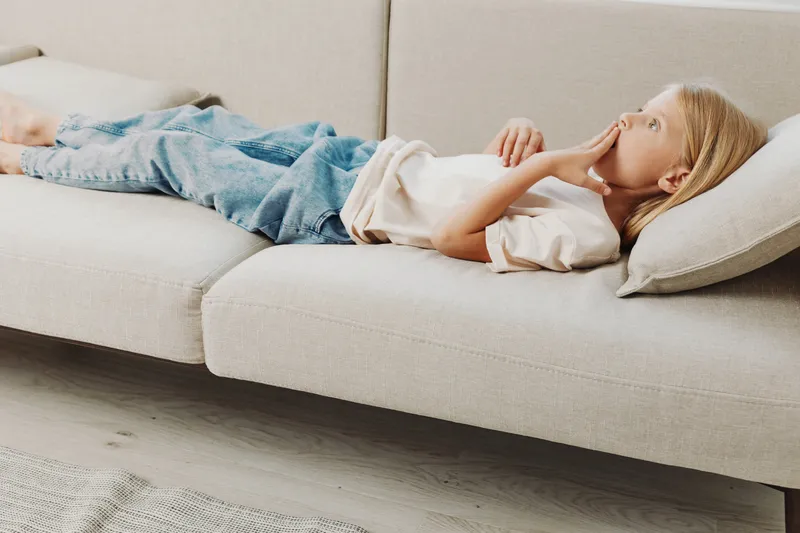 Young girl lies on beige sofa with pillow under head, wearing casual white t shirt and blue jeans. Concept of relaxation, childhood, home comfort and peaceful rest in modern living room.