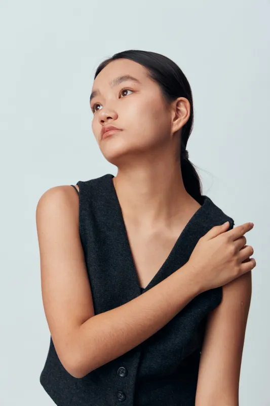 Young woman with long dark hair tied back posing in black sleeveless vest looking sideways on plain light background with thoughtful expression and crossed arm over chest.