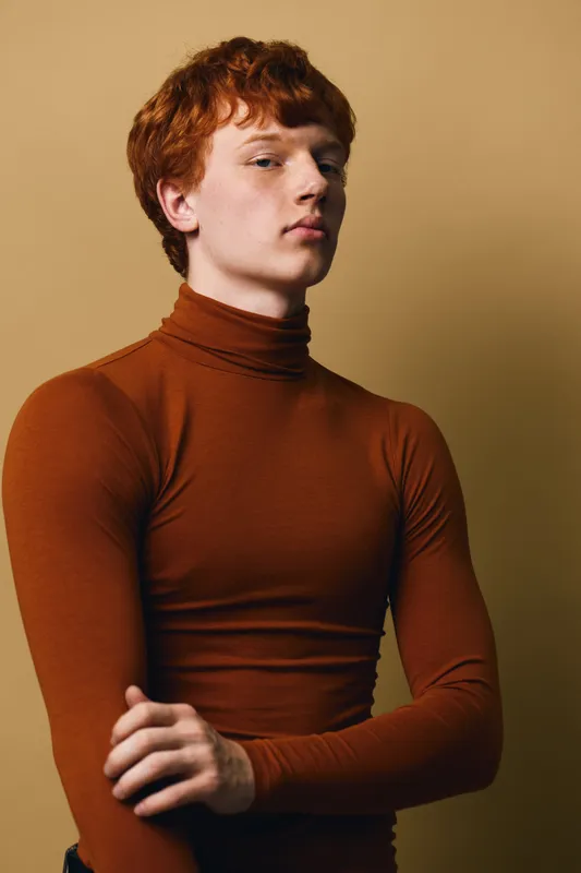 Young man with red hair wearing brown turtleneck sweater posing on beige background with confident expression in studio portrait fashion and style concept.