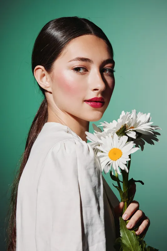 Portrait of a young woman with glossy hair holding white daisies. She wears a white shirt and poses against a green background. Concept for beauty creative fashion and elegance.