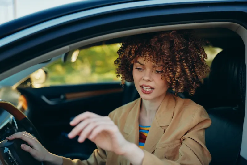 Cheerful woman inside a car, driver focused on the road, steering and wheel in frame, interior glow from sunlight, blazer and striped top, curly hair, warm candid lifestyle moment