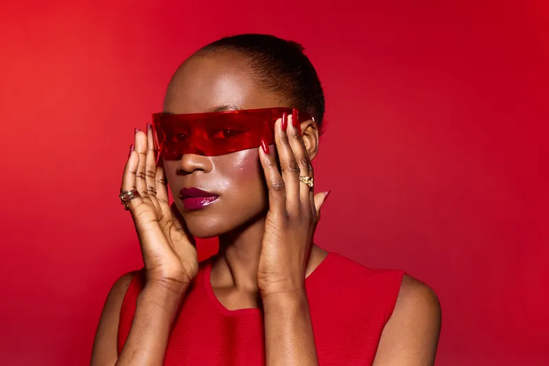 African American woman with dark skin wearing red dress and red transparent glasses poses against red background. Concept of fashion, style, beauty, and confident expression.