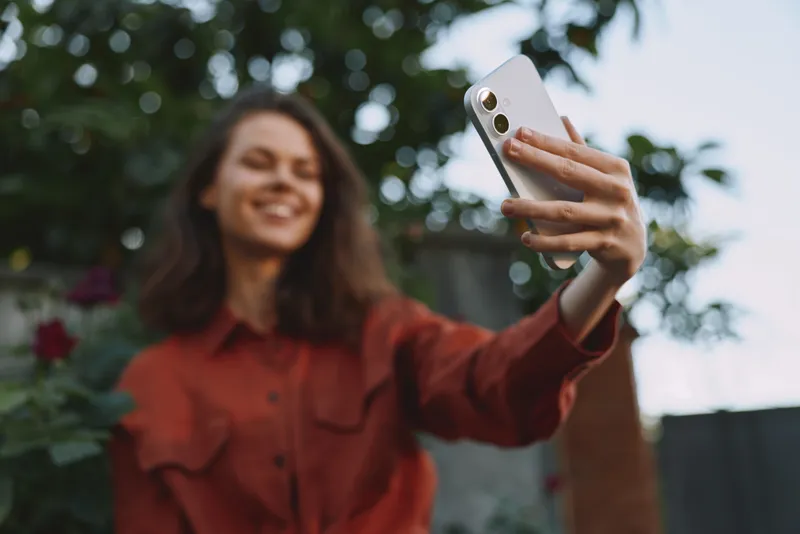 A young woman with shoulder length hair takes a selfie outdoors in a rust colored shirt, smiling as she holds a smartphone toward herself, surrounded by greenery and warm daylight