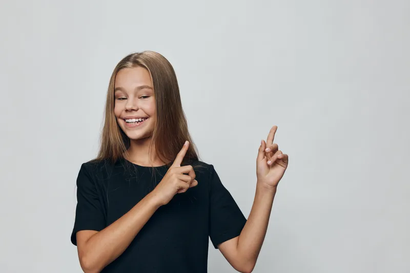 Girl, smile and happy with long hair wearing black t shirt pointing with fingers on gray background for positive expression and youth emotion in casual style portrait