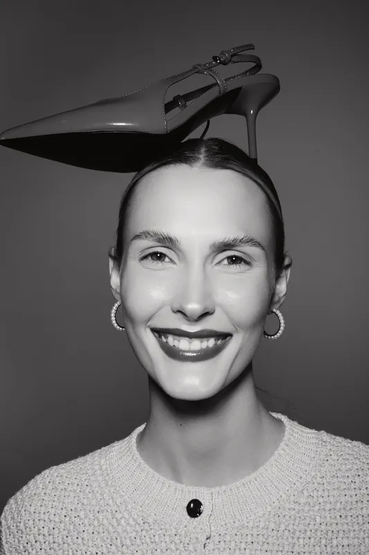 Smiling Caucasian woman with pale skin balances high heel shoe on her head. Studio portrait in black and white with joyful facial expression and casual knitwear. Concept of fashion fun.