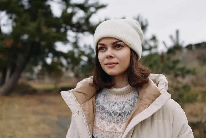 Winter portrait of a woman in a beige coat and knit sweater, wearing a beanie, outdoors among trees and open landscape.
