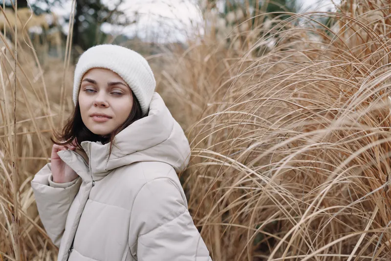 Woman in a light winter coat and beanie stands in a field of tall dried grass, calm expression, outdoor portrait with soft neutral tones and seasonal mood.