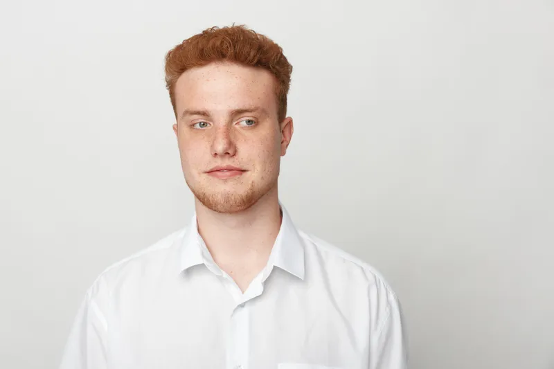 Young redhead man in white shirt looks sideways with thoughtful expression. Studio portrait on plain light background. Concept of contemplation, casual style, and natural appearance.