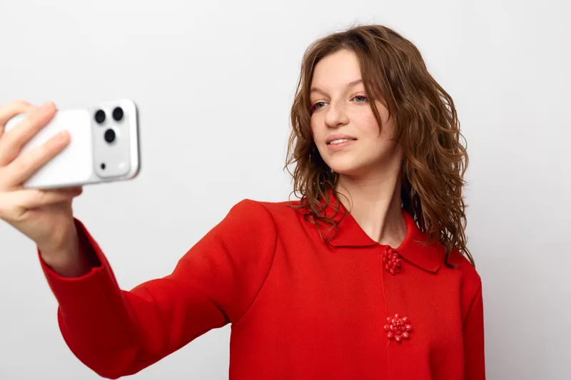 Young woman with curly hair wearing red sweater takes selfie with smartphone on white background. Casual style, technology use, and social media concept with natural expression and soft lighting.