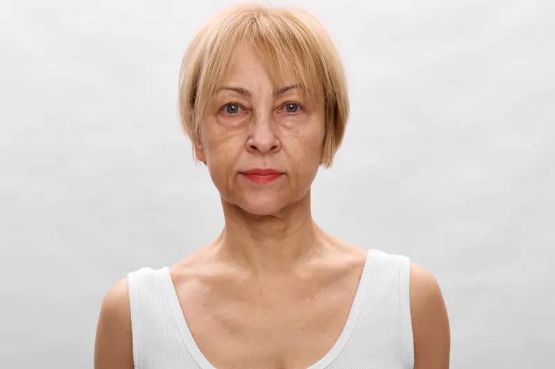 Senior woman portrait, neutral serious expression, pale blond hair, looking at camera in studio light, soft focus plain gray background, calm mood, introspection, quietly