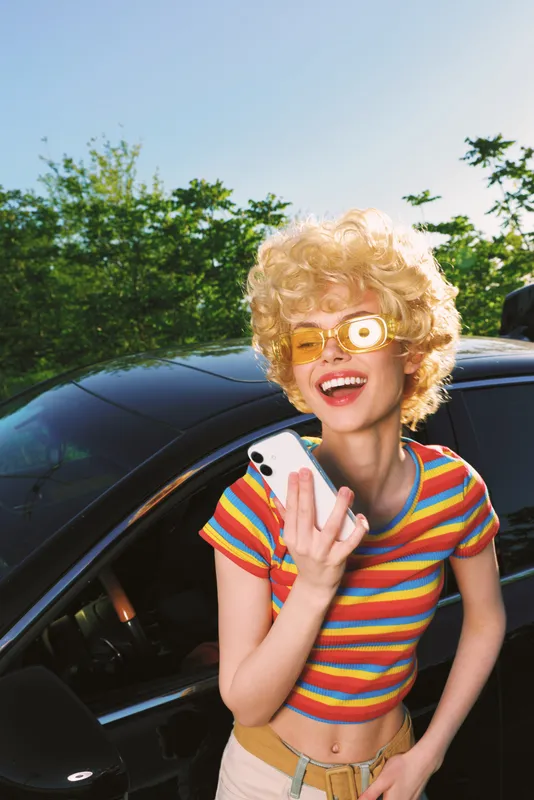 A woman in a striped shirt and retro sunglasses stands beside a car, capturing a selfie with bright sunny light. A candid lifestyle portrait with vintage flair and cheerful energy.