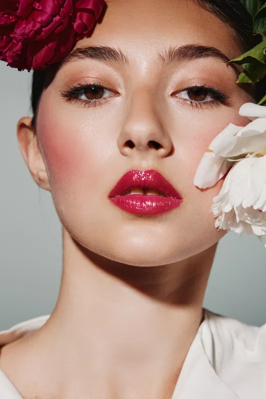 Beautiful woman with flawless skin and glossy red lips wears floral headpiece while holding white flower. Studio close-up shot features elegant fashion beauty creative concept.