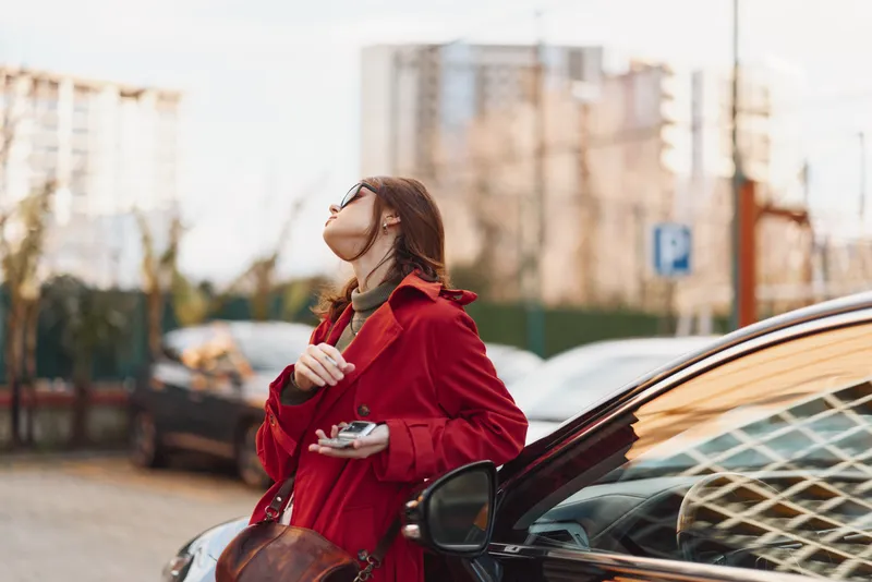 A woman in a red coat stands beside a black car on a city street holding a phone and looking up thoughtfully. Urban lifestyle and autumn fashion concept.