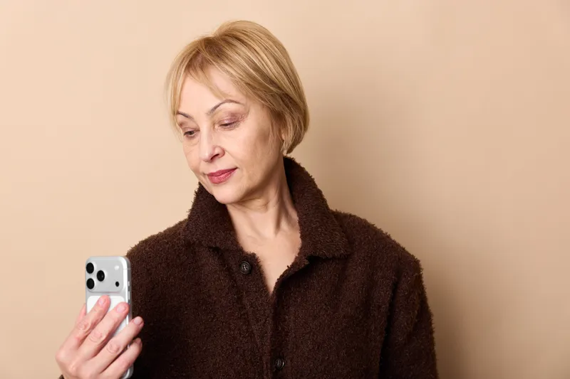 Elderly woman portrait holding smartphone, looking down with thoughtful expression, home communication using mobile technology, close up on face and hands, calm mood