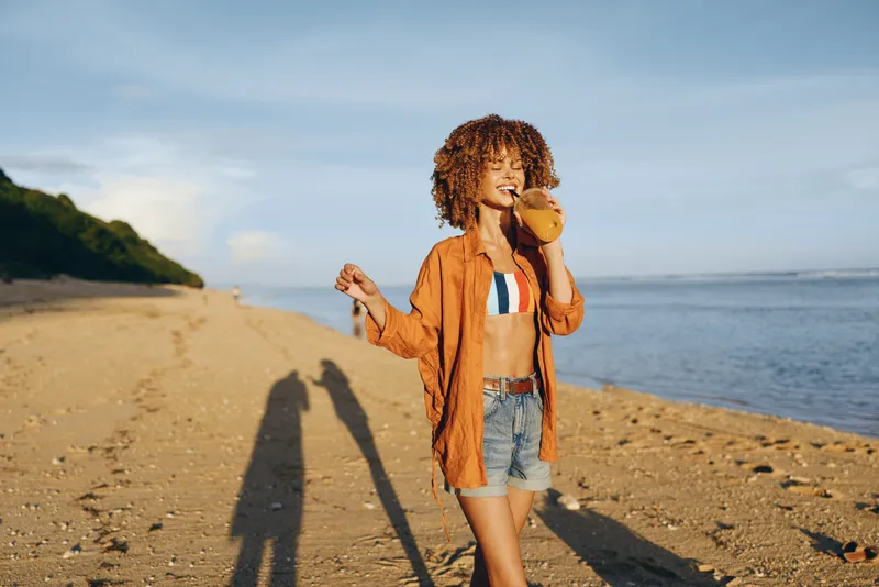 Woman with curly hair wearing orange jacket and denim shorts walks along sandy beach by the ocean in summer sunlight, relaxed vacation mood, long shadow on ground near cliffs horizon