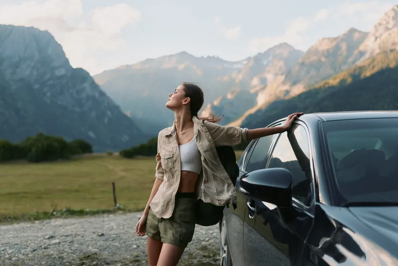 Young woman in casual clothes leans on a car and enjoys fresh mountain air with closed eyes during a peaceful outdoor moment in nature.