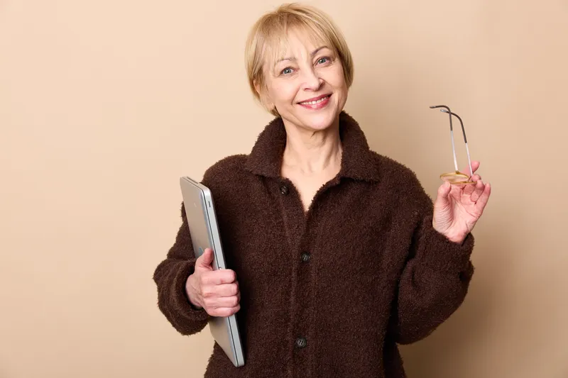 Smiling elderly woman portrait, holding smartphone in one hand and glasses in the other, relaxed casual outfit, beige studio background, ready for communication technology, read
