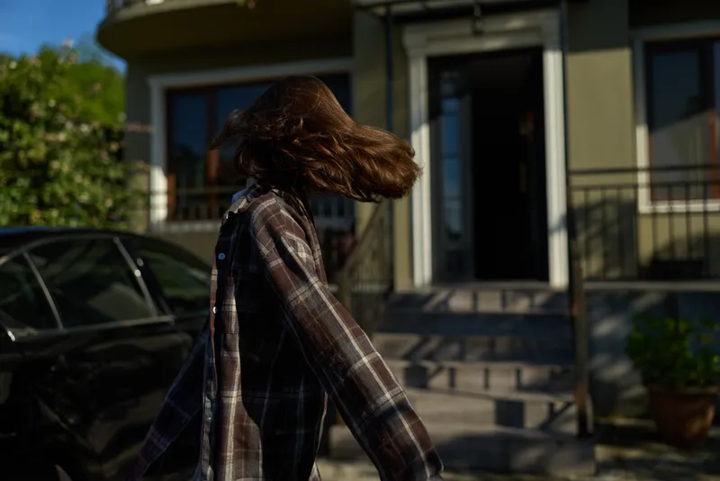 A young woman in a plaid shirt walks away from the camera along a sunny residential street, hair swept by the wind, capturing a candid everyday lifestyle moment