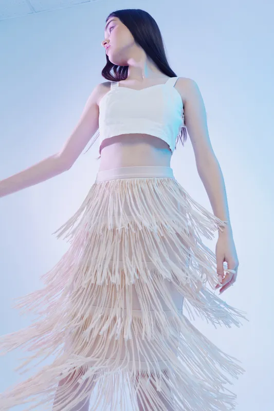 Young woman in a white crop top and fringed skirt poses gracefully against a soft blue background. Fashion and beauty concept with elegant movement and modern style.