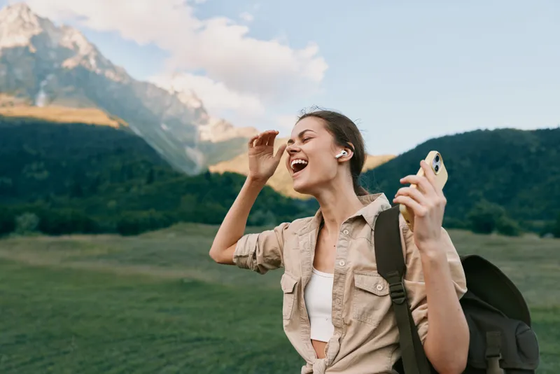 Young woman with earbuds enjoys music outdoors in a mountainous landscape while holding a smartphone and smiling with eyes closed in a carefree moment.