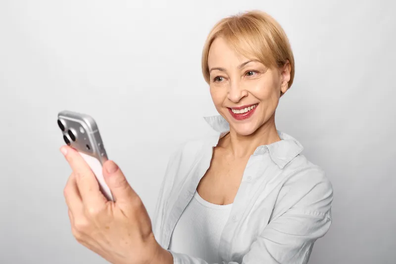 Woman smiling using smartphone, elderly portrait indoors, looking at camera, bright white studio background, relaxed hand gesture, communication technology