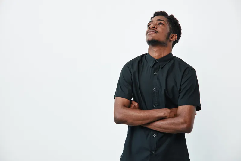Young black man with dark skin wearing black shirt standing with arms crossed looking up on white background. Confident male portrait with casual style and thoughtful expression.