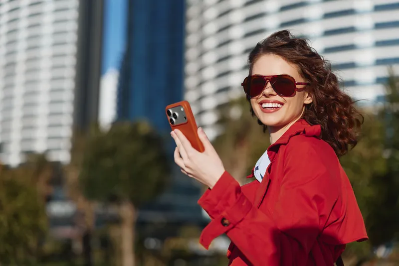 Cheerful woman outdoors, wearing a red jacket and sunglasses, smiling as she holds a phone for a selfie, urban background, sunny day, relaxed mood