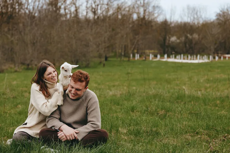 Smiling couple sitting on grass with a friendly goat kid in a rural field. Casual sweaters, relaxed mood, outdoor lifestyle, connection and playfulness, warm natural light. Warm, cozy vibes.