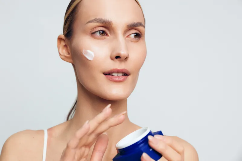 Young woman applies facial cream on cheek holding blue jar skincare product with gentle expression isolated on white background for beauty care concept.