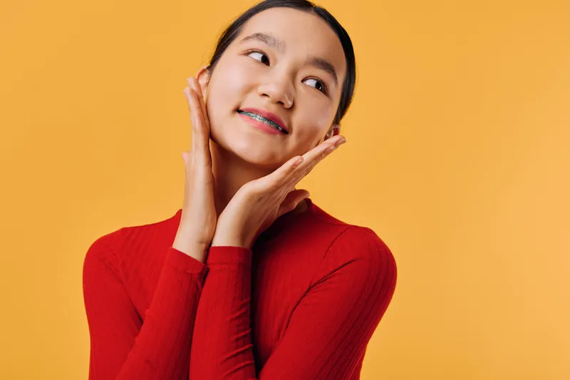 Young woman with braces smiling and posing with hands near face on yellow background. Portrait of happy female, beauty, fashion and casual style with positive emotion and confident look.