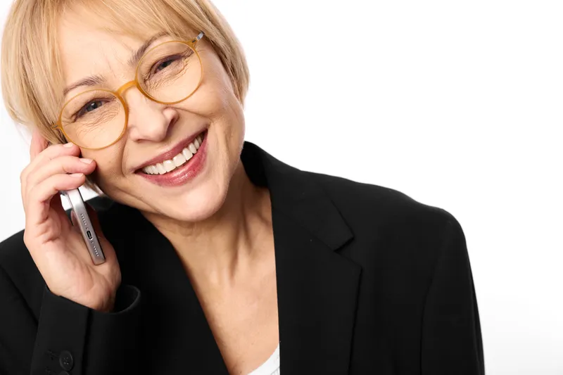 Woman smile blonde glasses phone call communication business portrait closeup, cheerful adult woman in blazer holding phone to ear, looking at camera, white background, soft studio lighting, friendly