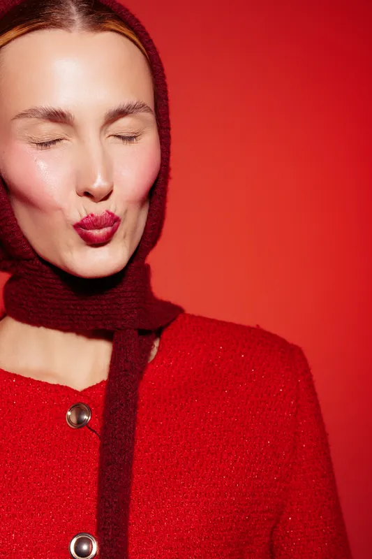 Young woman with closed eyes wearing red sweater and scarf blows a kiss on red background. Concept of affection, warmth, and cozy fashion in studio lighting.