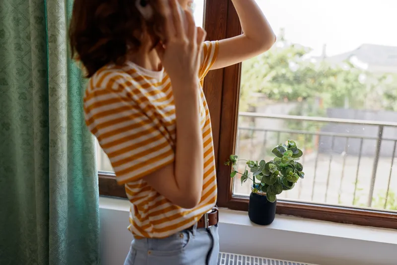 Woman indoors by a window, wearing a striped shirt, looking outside with a calm contemplative expression, daylight highlights a cozy home moment with a plant nearby
