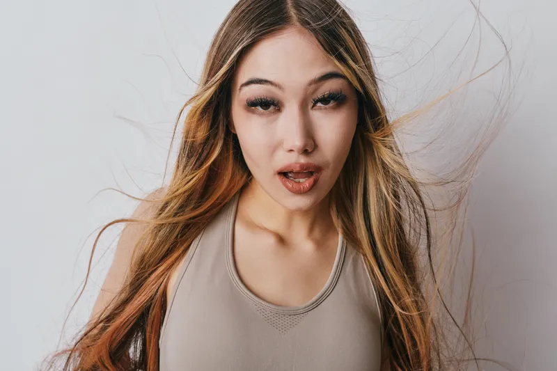 Young woman with long hair and neutral expression posing on light background, natural makeup, casual style with beige top and flowing hair movement effect.