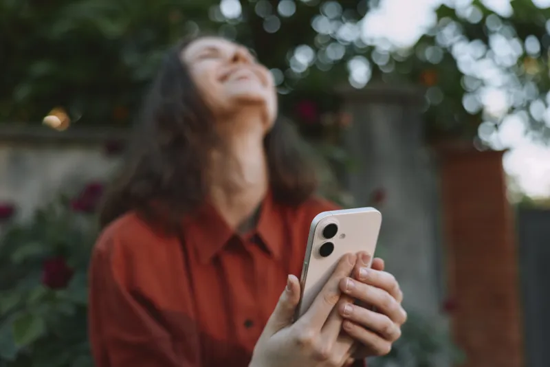 Smiling woman in a red shirt holding a smartphone outdoors, looking up with joy. Casual lifestyle scene with natural light, garden backdrop, and relaxed mood. Warm vibe