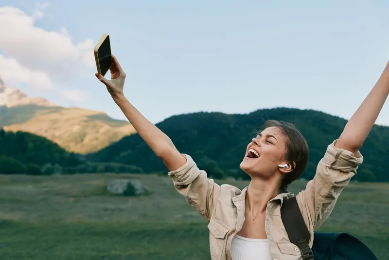 A joyful woman outdoors raises her arms and holds a smartphone, enjoying nature and fresh air in a mountainous landscape. Concept for lifestyle and happiness.