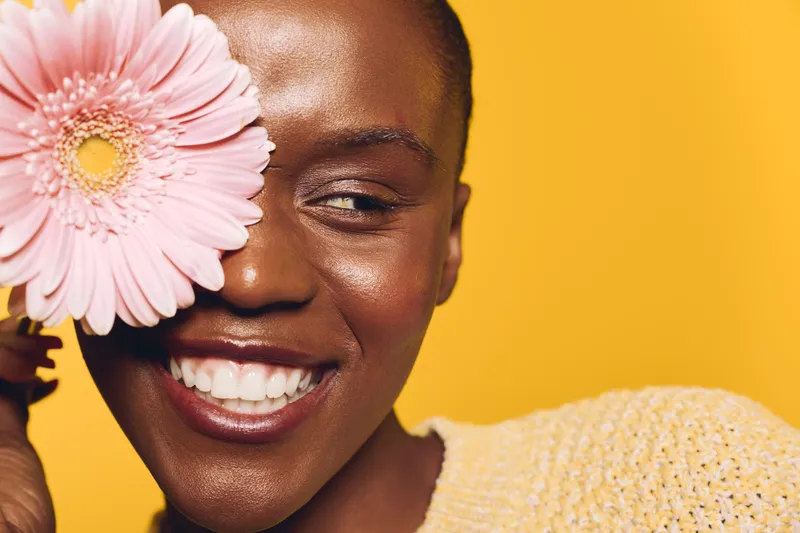 Close-up of happy African woman with dark skin smiling and holding pink flower near eye. Bright yellow background and cozy sweater create warm and cheerful mood for beauty and nature concept.