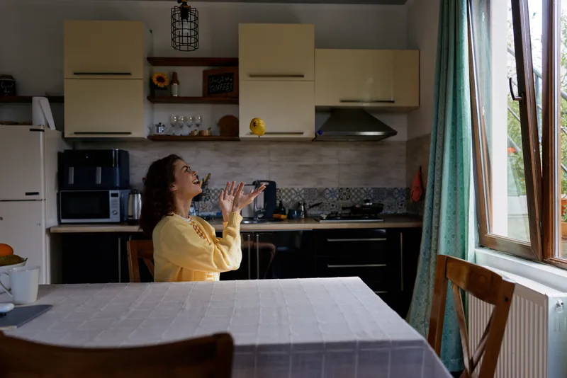 Cheerful woman in a cozy kitchen, wearing a yellow sweater, looking up with hands raised, sunlit scene, casual lifestyle moment, warm colors, tidy surroundings, candid atmosphere