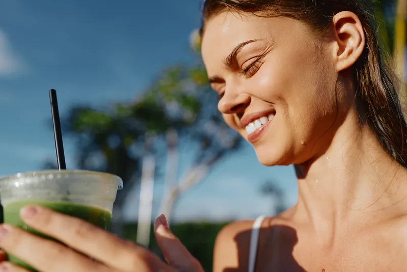 Cheerful woman outdoors, smiling while holding a smoothie, sunlit moment, relaxed posture, natural colors, garden backdrop, healthy lifestyle and refreshing vibe in bright daylight.