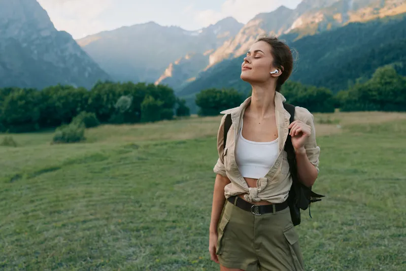 A young woman in casual summer clothes enjoys a peaceful moment outdoors with mountains and green fields in the background. Concept of lifestyle and nature relaxation.