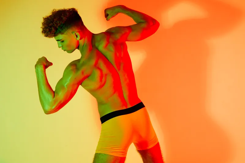 Muscular man posing shirtless in colorful lighting with flexed arms showing strength and fitness. Back view of athlete with defined muscles and curly hair in studio setting.