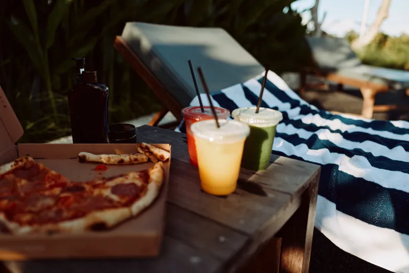 Outdoor picnic scene with pizza slices and colorful drinks on a wooden table, warm sunlight and relaxed summer mood, casual lifestyle, fresh food, social moment, leisure and happiness.