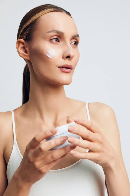 A young woman applies facial moisturizer in a bright studio, her concentrated expression capturing a personal skincare moment with clean lighting and a minimal lifestyle focused on wellness and daily